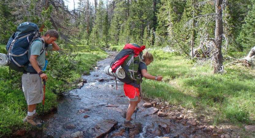 A person wearing a backpack crosses a creek in ankle-deep water while another person watches